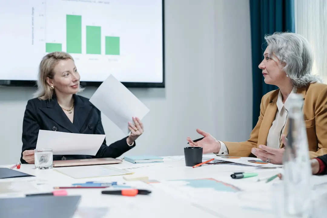 Due donne manager in azienda che, sedute alla scrivania, con una lavagna alla spalle e dei fogli in mano, discutono positivamente delle questioni aziendali | 4Humans